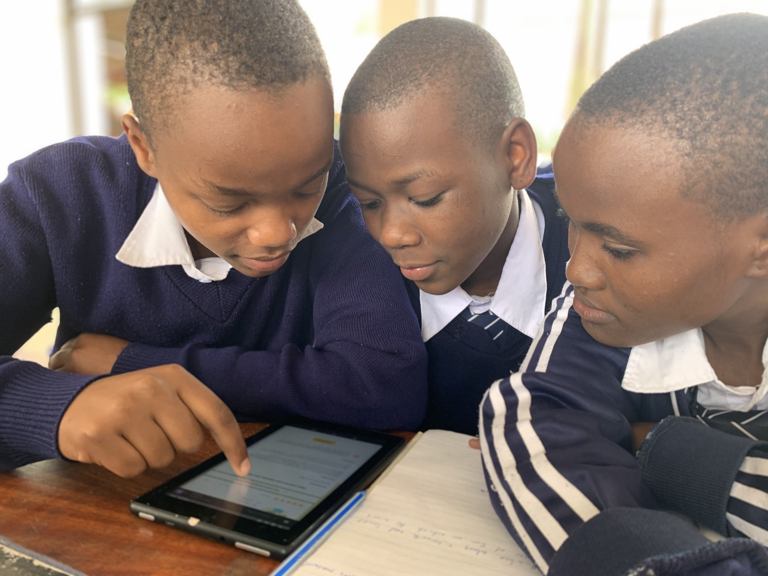 Three students gather around a tablet to read what's on the screen. The student on the left hand side points to something on the screen with his index finger.