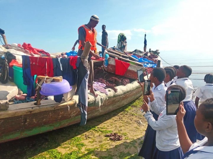Students from St. Monica Secondary School in Zanzibar gather around a fishing boat as a man on the boat shows them fishing gear. Some of the students use tablets to take pictures of the demonstration.