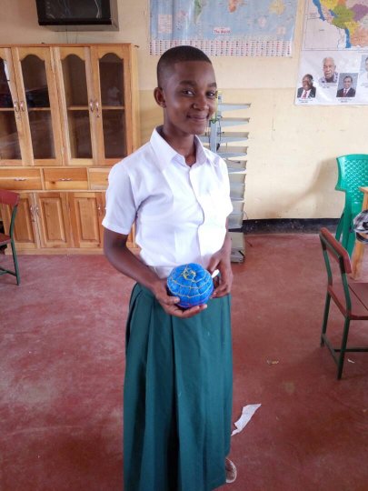 A young student smiles and presents her an object she designed to the camera. The object is a blue sphere with horizontal and vertical lines sewn into it.