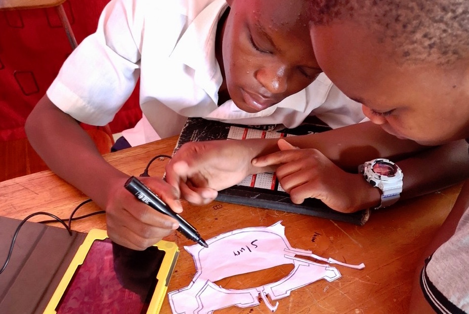 Two secondary school students collaborate on a project. One holds a marker and is writing on paper that has been cut out into a design.