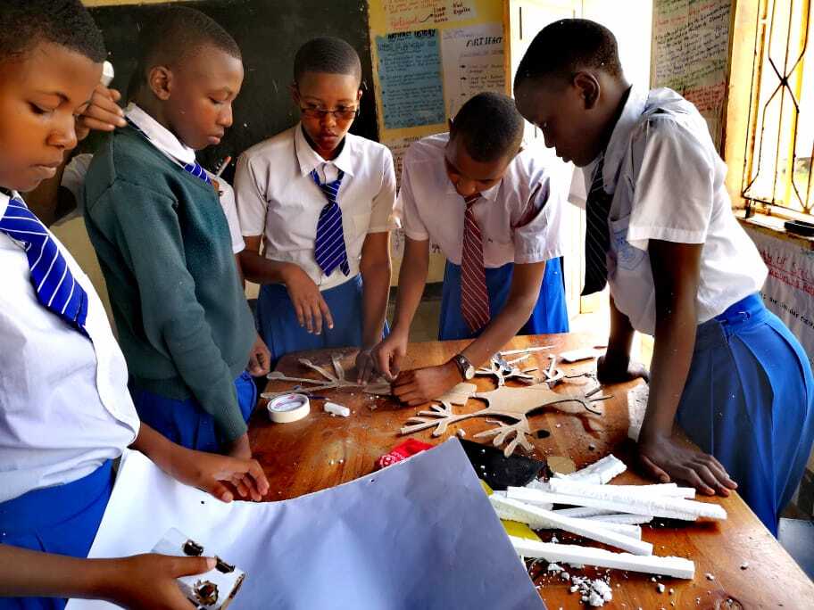 Four students appear deeply engaged in their work, assembling a a complex cardboard structure on a work table in a Quest Forward School in Tanzania.