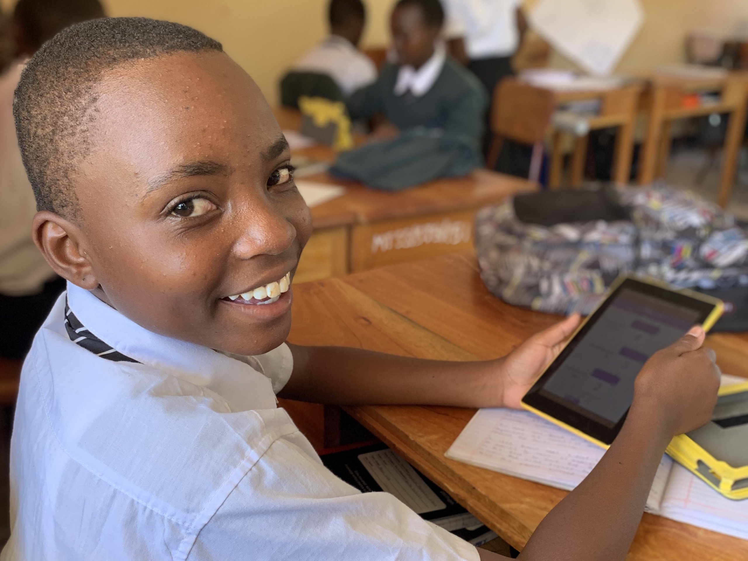 A student smiles brightly over his shoulder, while holding a tablet displaying the Quest app in his secondary school classroom in Tanzania.