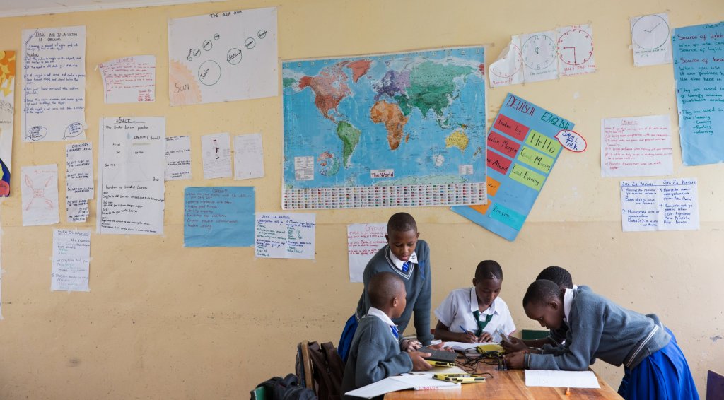 Student work covers the wall behind students working in a secondary school classroom in Tanzania.