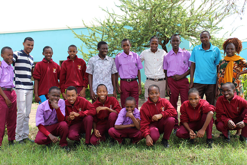 Students and staff at Arusha City Boys pose outside. The students wear red slacks and either a lavender or red shirt.