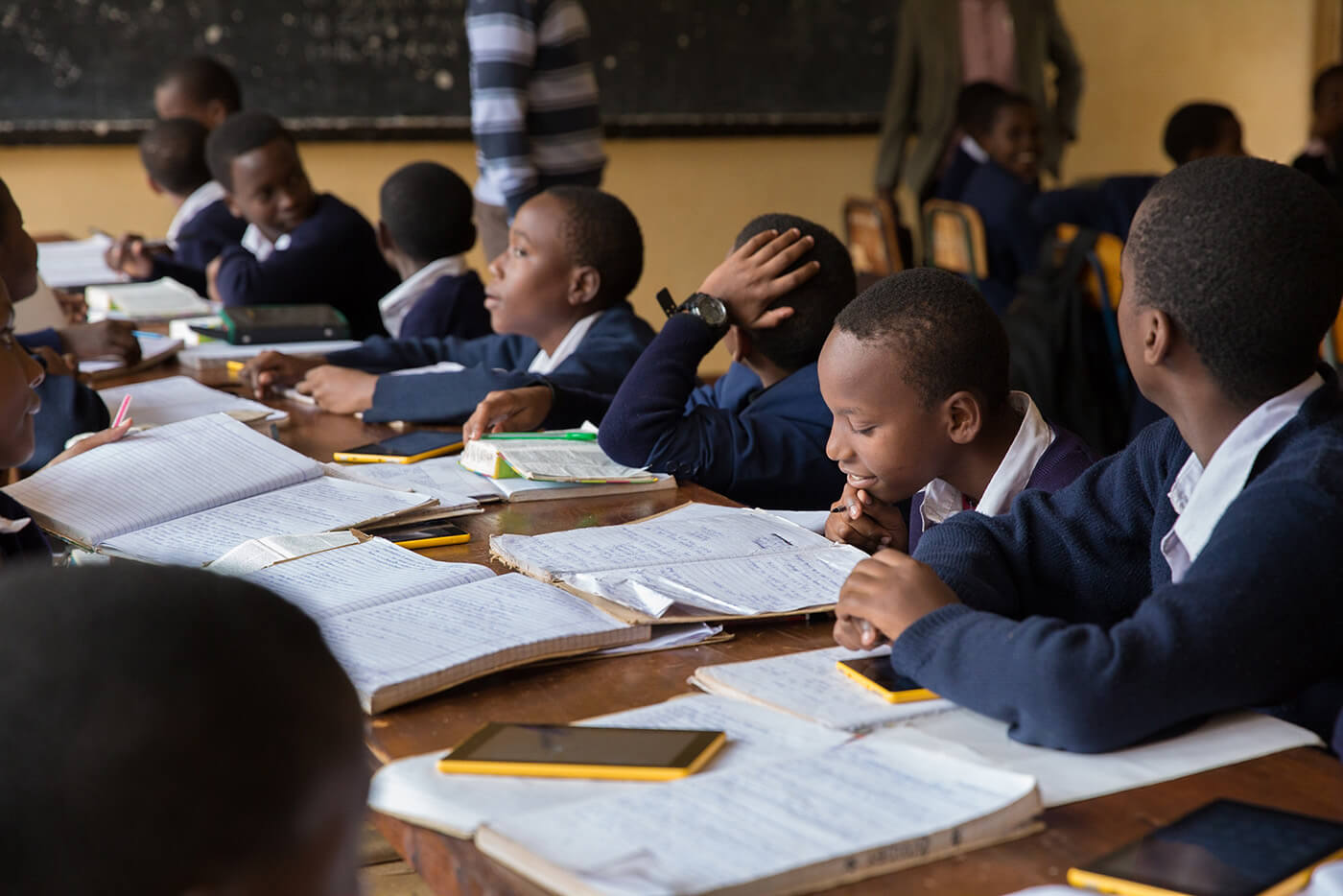 Students sit along a long table. In front of them are different learning materials such as course books, note books, and tablets.