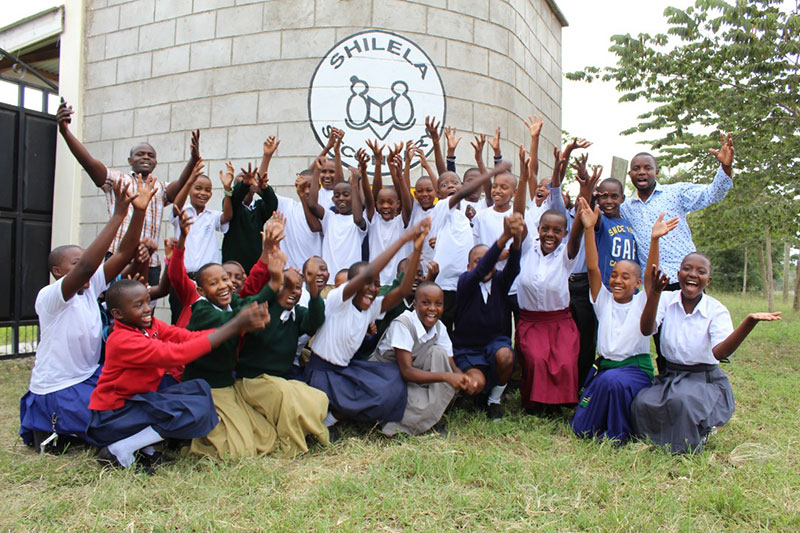 Students at Shilela Secondary School pose together outside the school, smiling and cheering with their arms extended.
